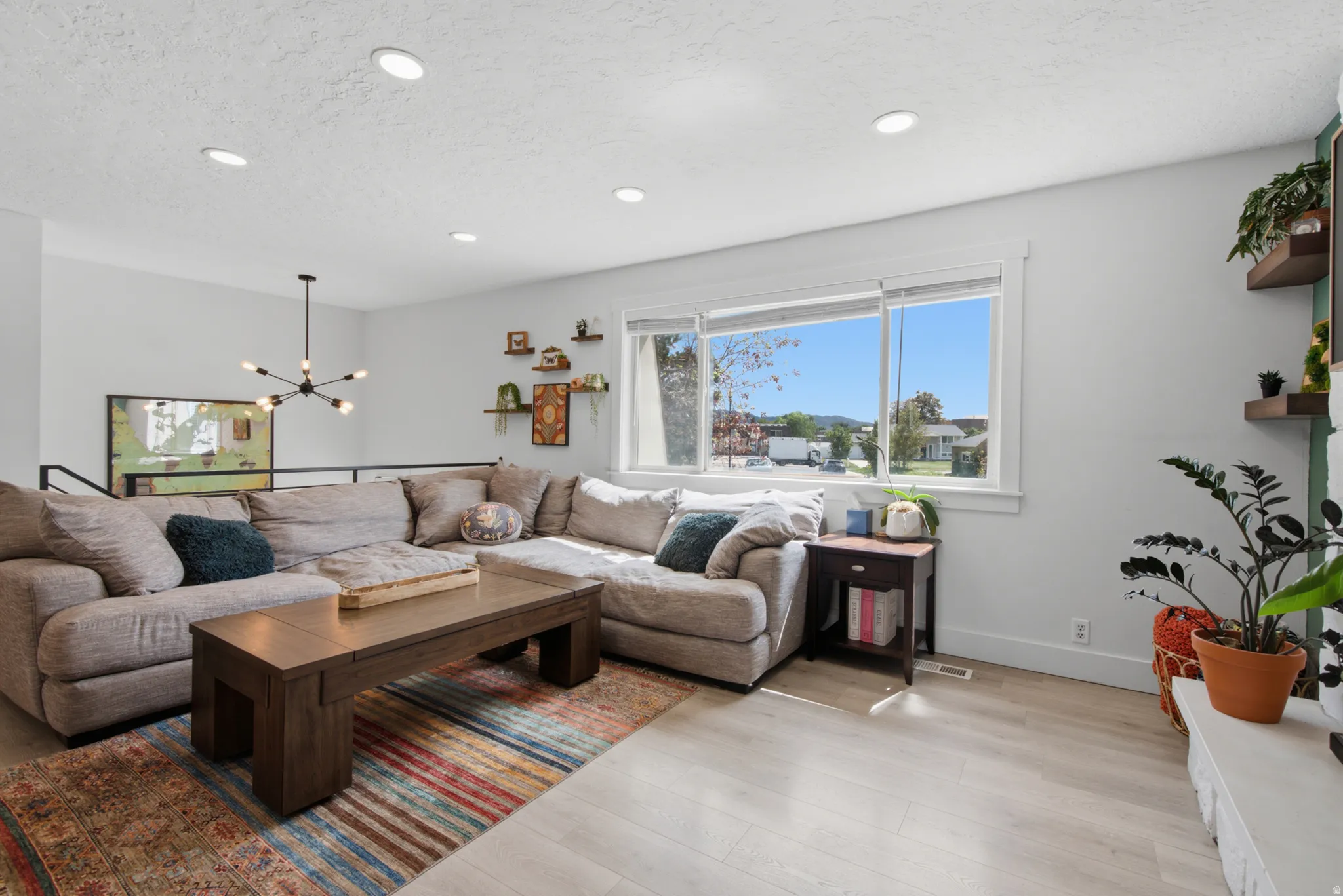 Recessed ceiling lighting, wood-finish flooring, large window with white trim, black metal railing, and a contemporary chandelier