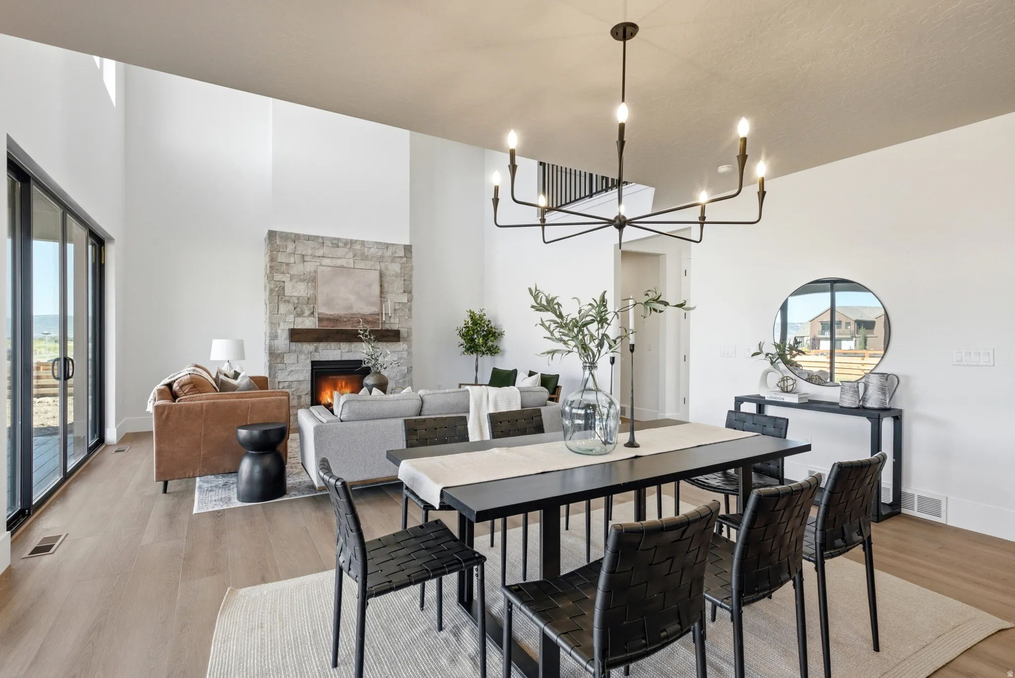High ceilings, wood-finish flooring, stone-clad fireplace with wood mantel, large sliding glass doors, and a modern chandelier