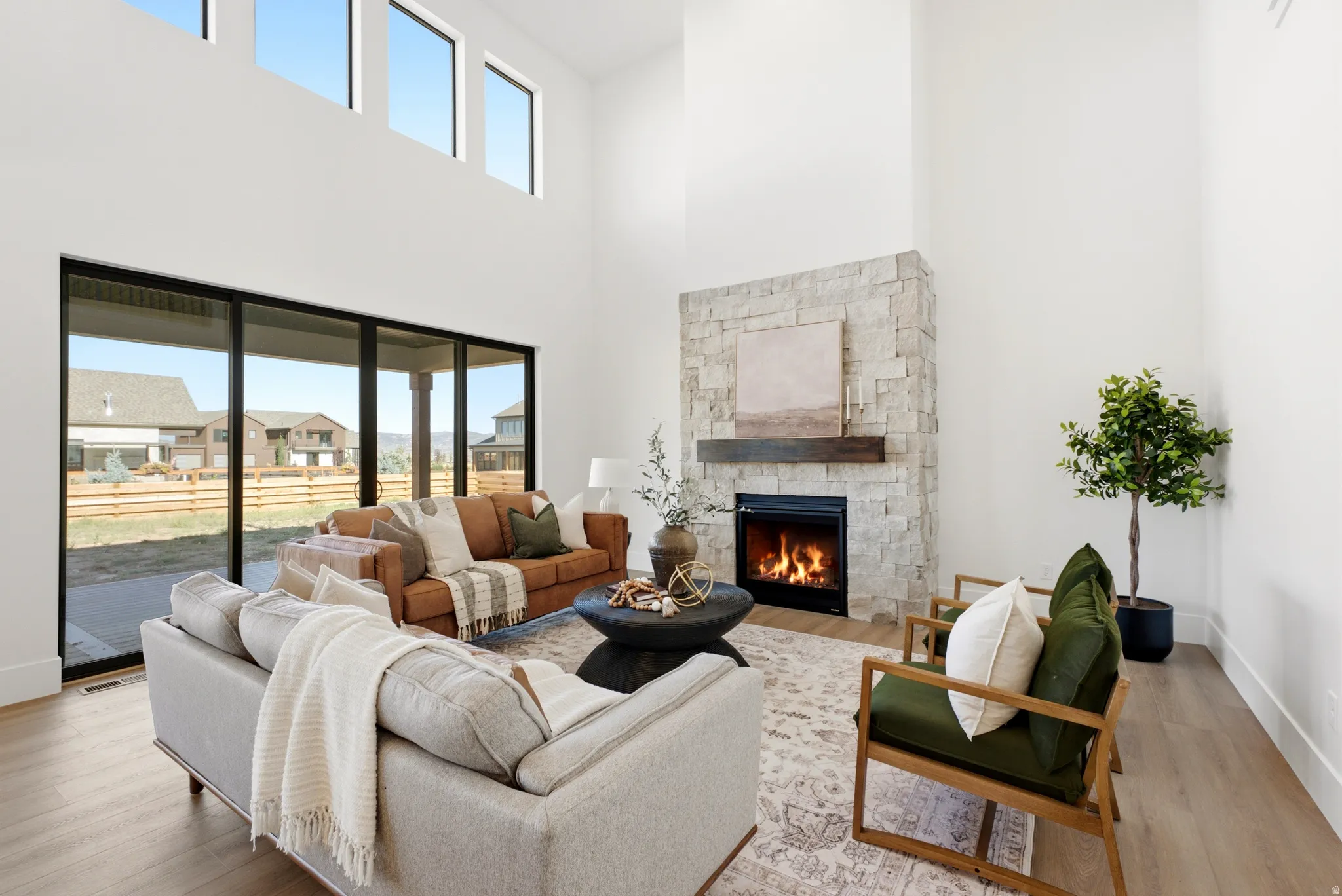 Soaring ceiling with high windows, full-height stone fireplace with a wood mantle, sliding glass doors to the exterior, and wood-finish flooring
