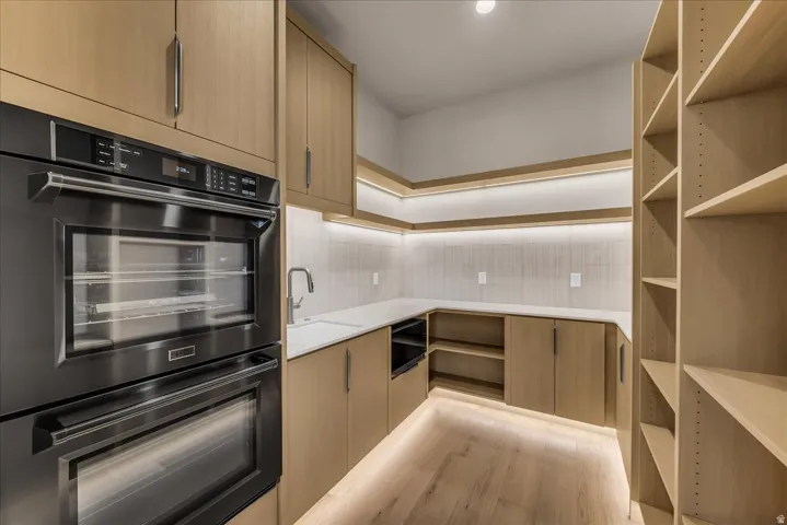 Built-in double ovens, light wood-finish cabinetry with linear hardware, wood-finish flooring, integrated under-cabinet lighting, and extensive open shelving