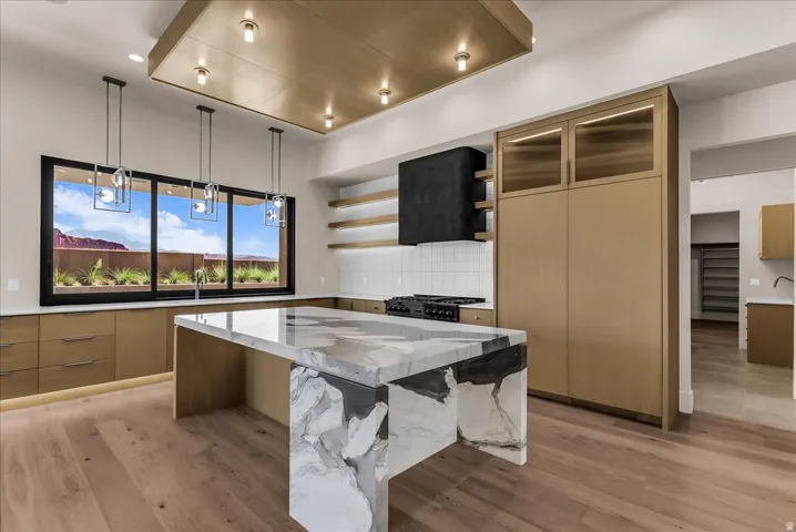 Waterfall island with high-contrast stone finish, wood-finish flooring, custom cabinetry, large window with integrated sink, and recessed ceiling with accent lighting