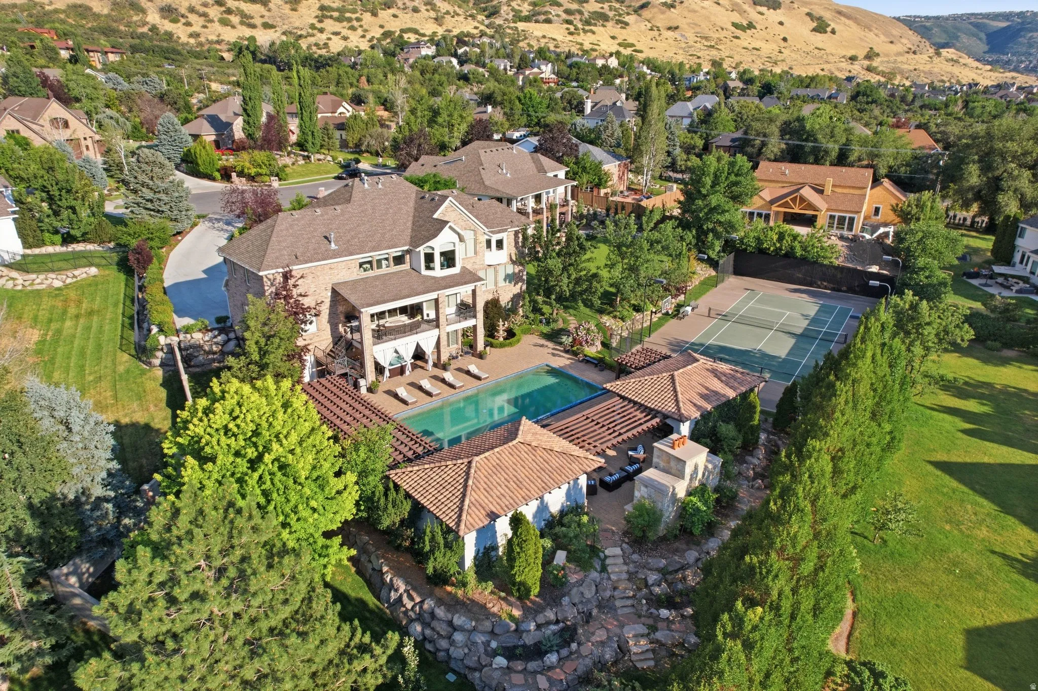 Expansive outdoor living area featuring a large swimming pool, a full-size tennis court, a covered patio, and a separate cabana with an outdoor fireplace