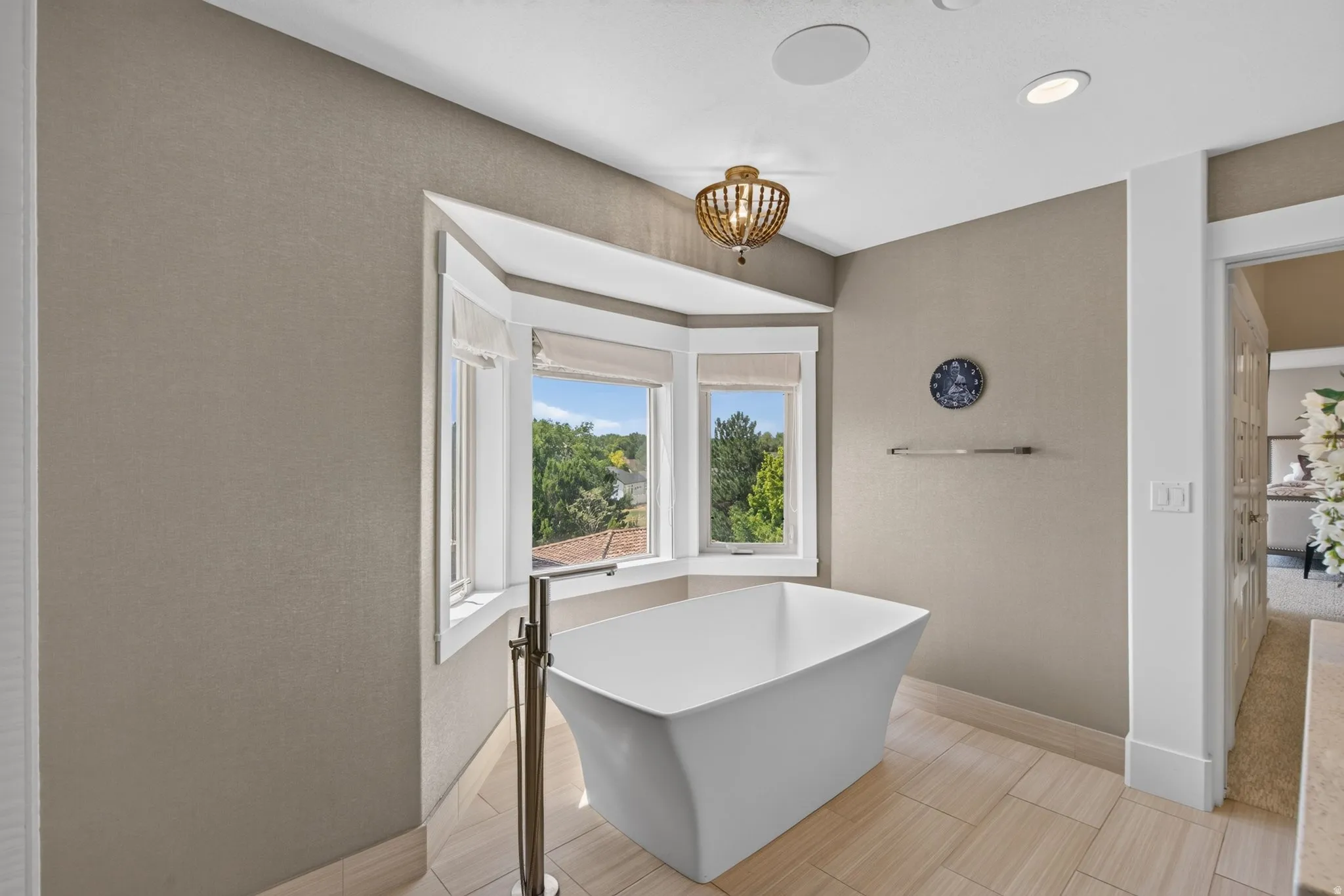 Freestanding soaking tub with floor-mounted brushed nickel faucet, bay window with window treatments, recessed lighting, and large format tile flooring