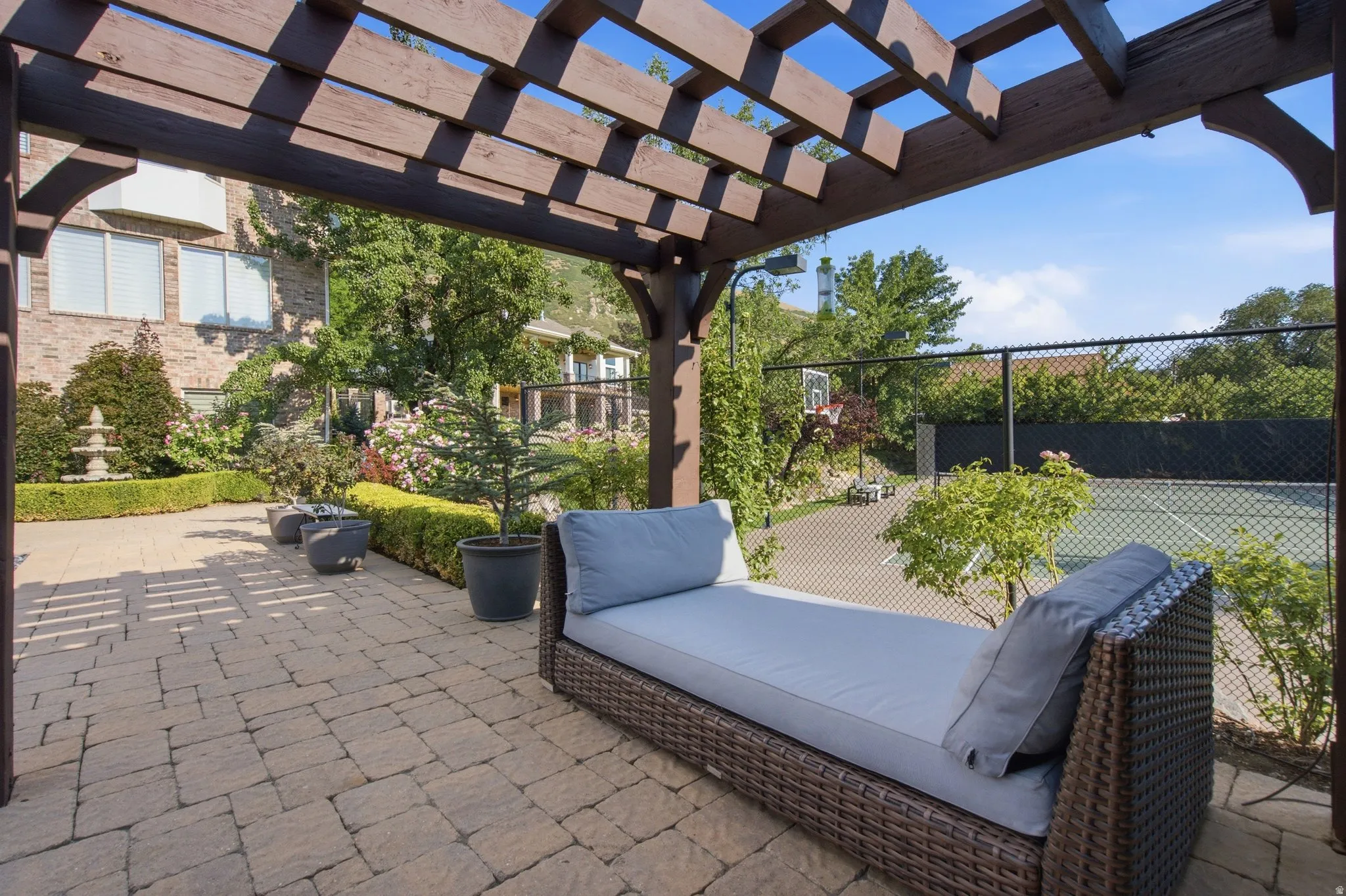 Expansive paver patio with a wood pergola, featuring a tiered fountain, lush landscaping, and a fenced sports court