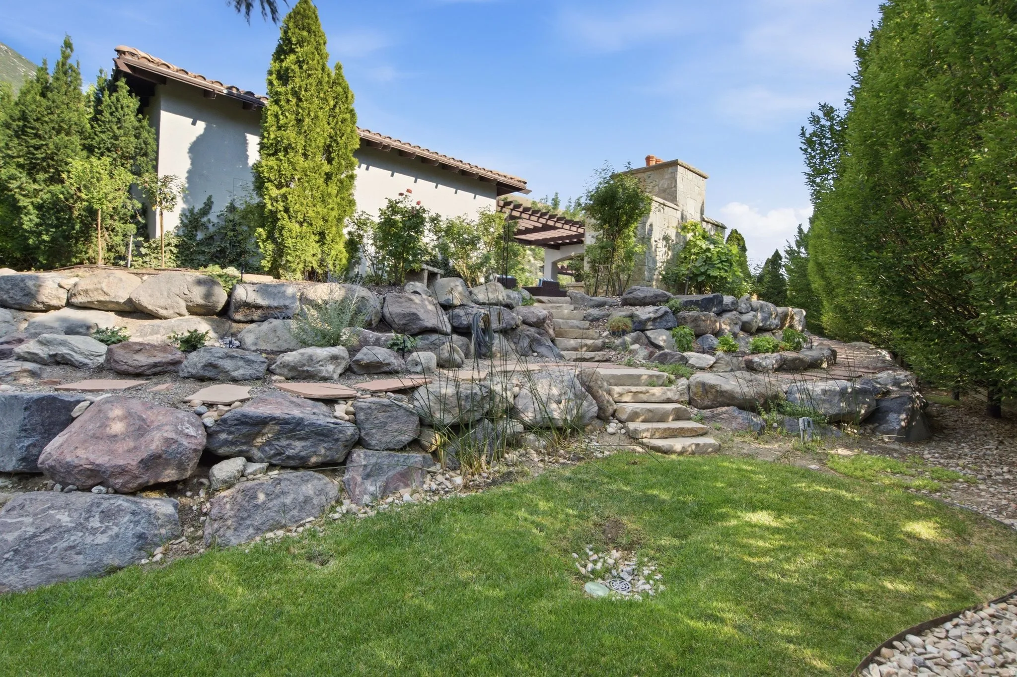Terraced landscape with natural stone retaining walls, stone steps, a pergola, and mature trees