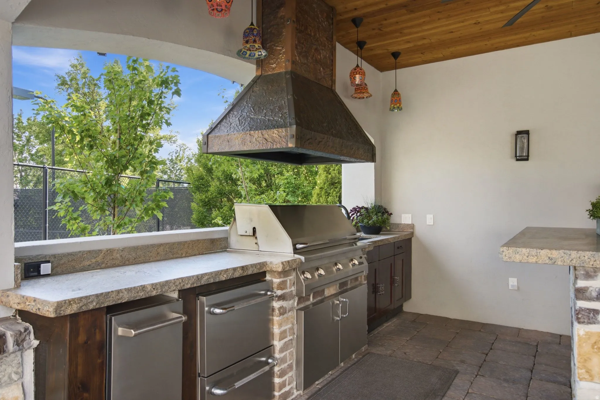 Outdoor kitchen featuring a built-in grill, stone countertops, stainless steel appliances, brick pillar accents, and a wood-panel ceiling