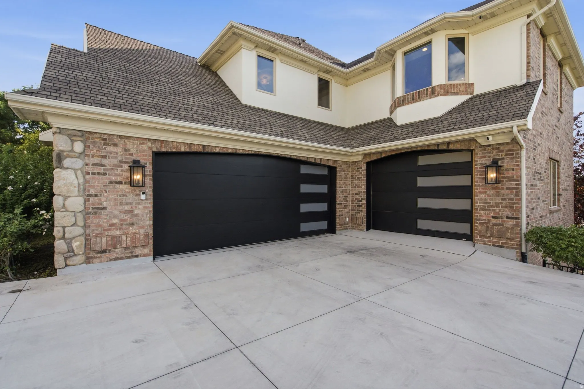 Two modern garage doors, brick and stone exterior detailing, stucco accents, and traditional outdoor lighting fixtures