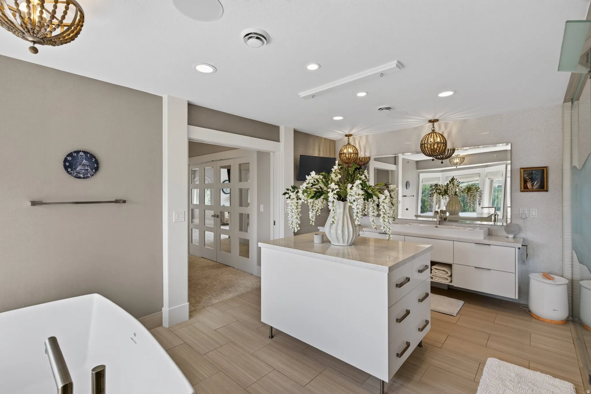 Freestanding soaking tub, dual vanity with undermount sinks, central island with storage, pendant lighting fixtures, and light wood-finish flooring