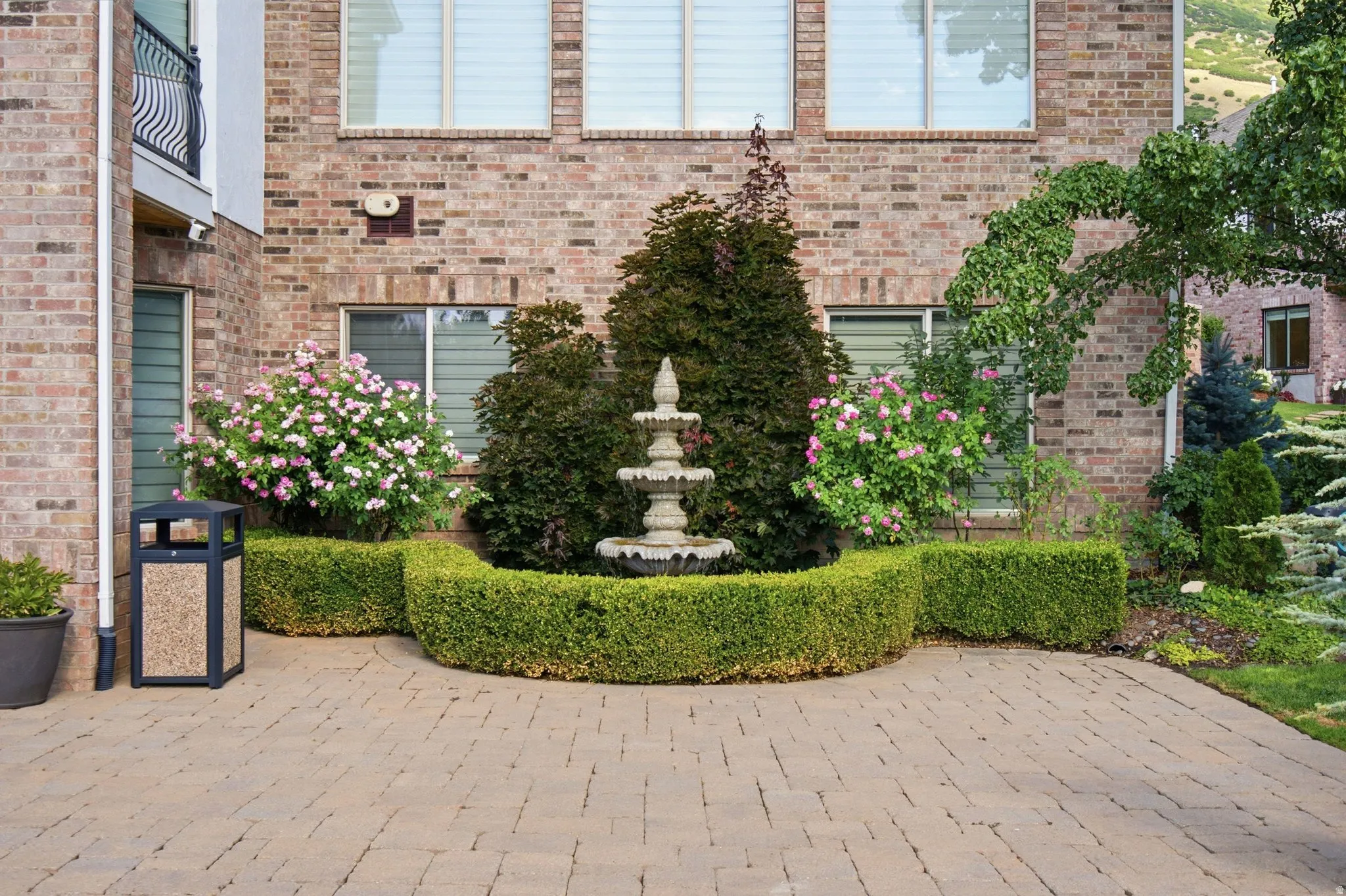 Brick exterior featuring multiple windows, a stone tiered fountain, manicured hedges, and flowering shrubs
