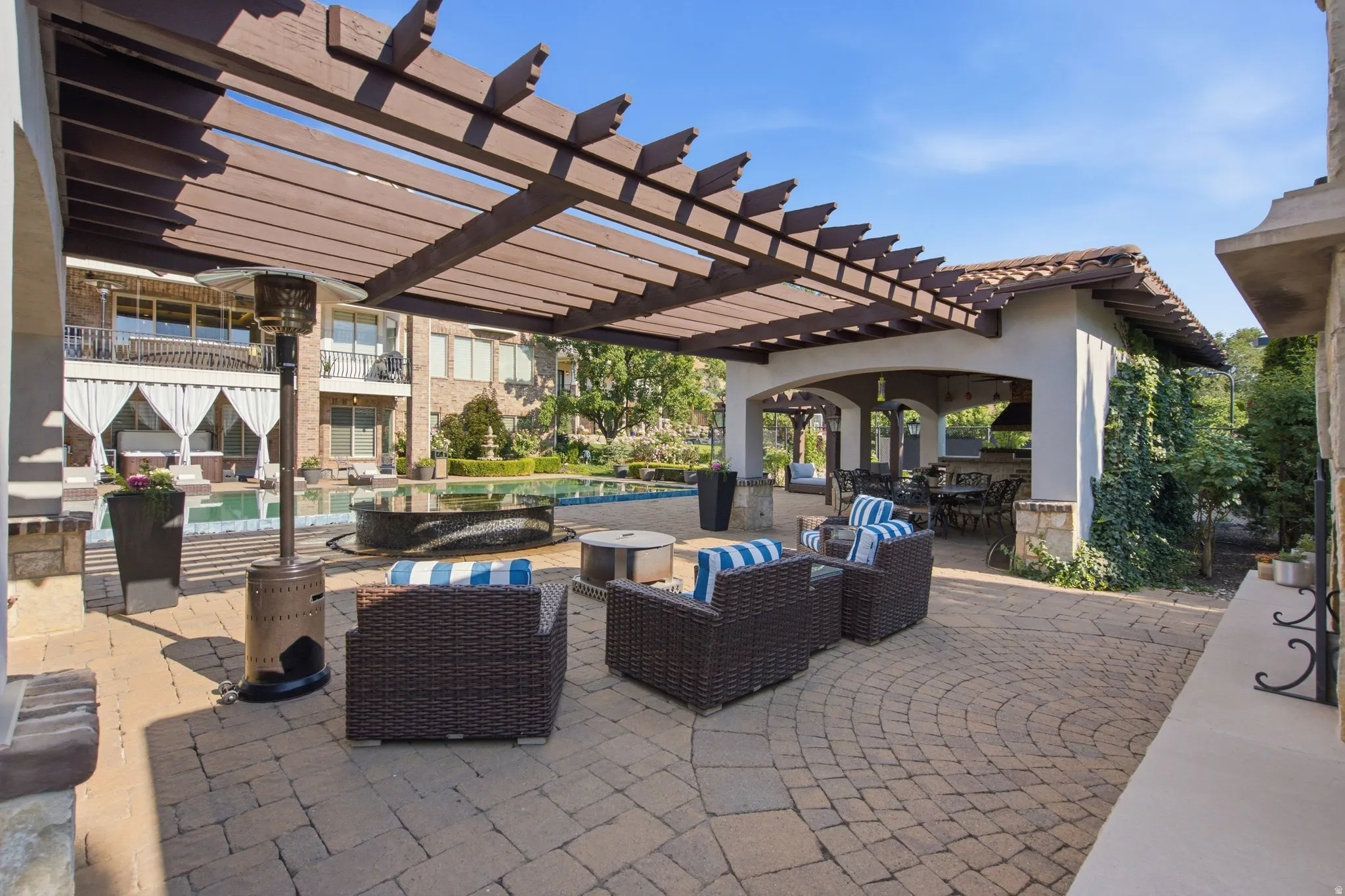 Expansive paver patio with a wood pergola, integrated fire feature, and a dedicated outdoor kitchen structure with a tile roof