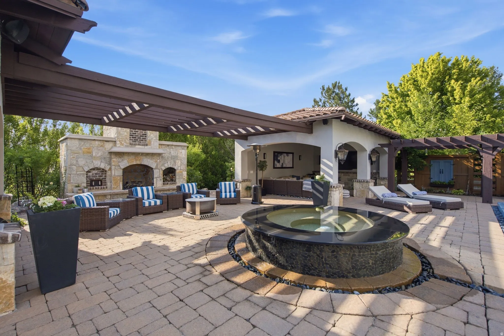 Expansive paver patio featuring a stone fireplace, pergola, built-in outdoor kitchen, and a circular water feature