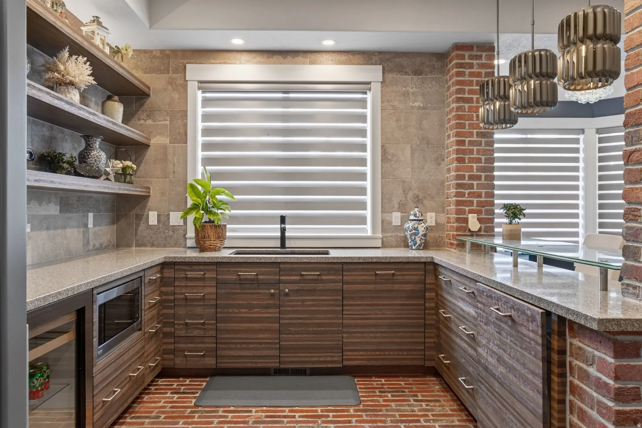 Rich wood-finish cabinetry, light-toned countertops, integrated microwave, beverage refrigerator, and exposed brick accents