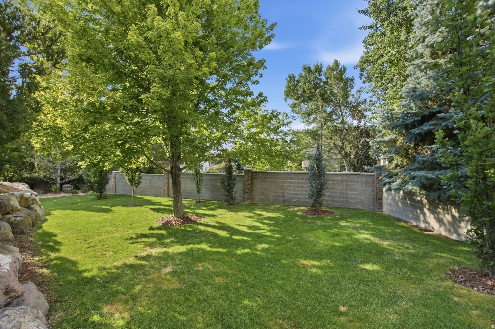 Expansive lawn area with mature trees, a retaining wall, and a block fence featuring stone accents