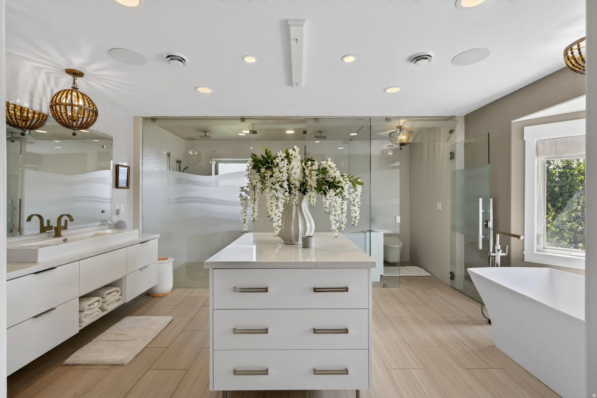 Dual floating vanity with integrated rectangular sinks, a freestanding soaking tub, a spacious glass-enclosed shower, a central island with a stone-finish countertop, and recessed lighting