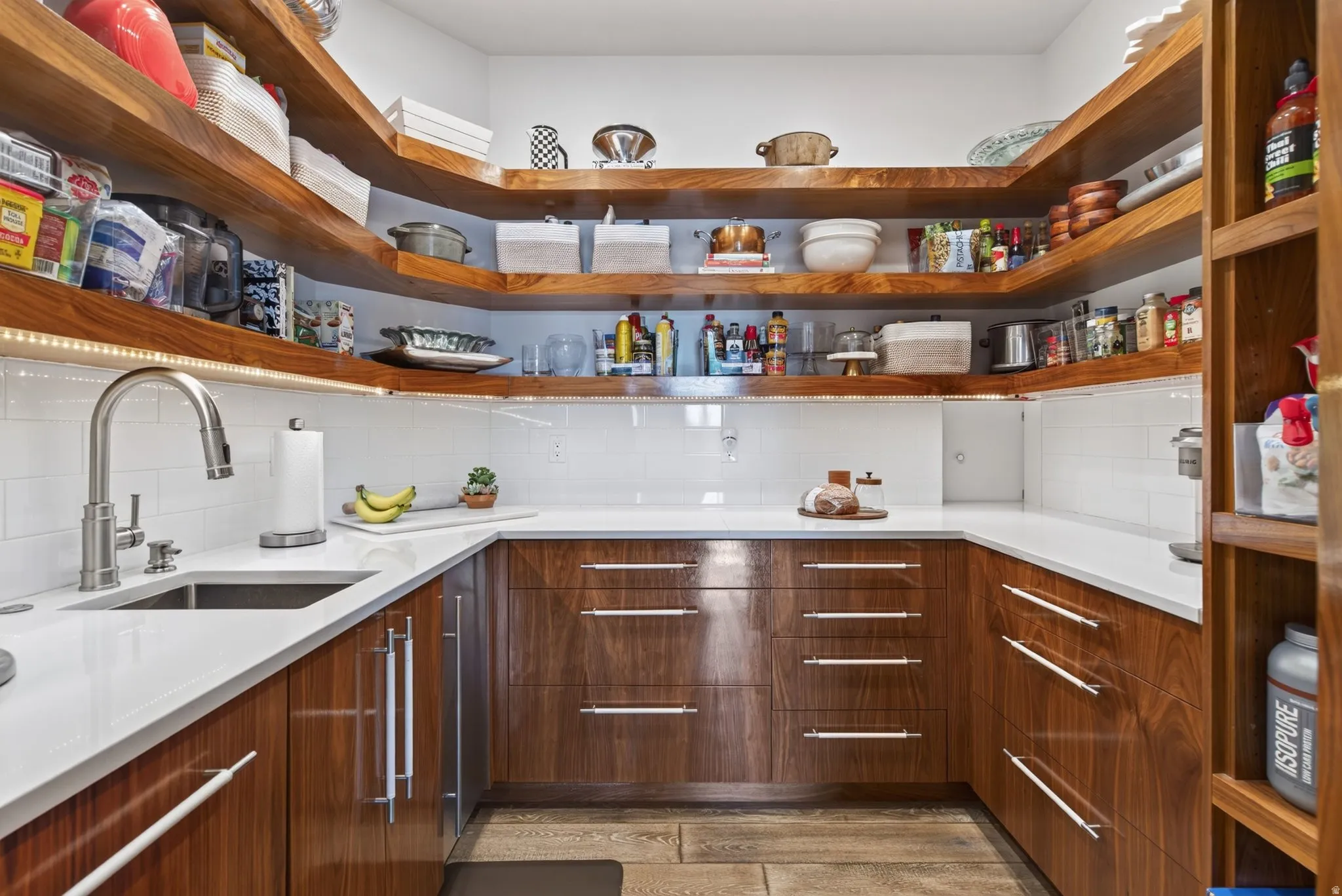 U-shaped pantry featuring wood-finish cabinetry, white countertops, subway tile backsplash, a stainless steel sink with a gooseneck faucet, and floating wood-finish shelving with integrated lighting