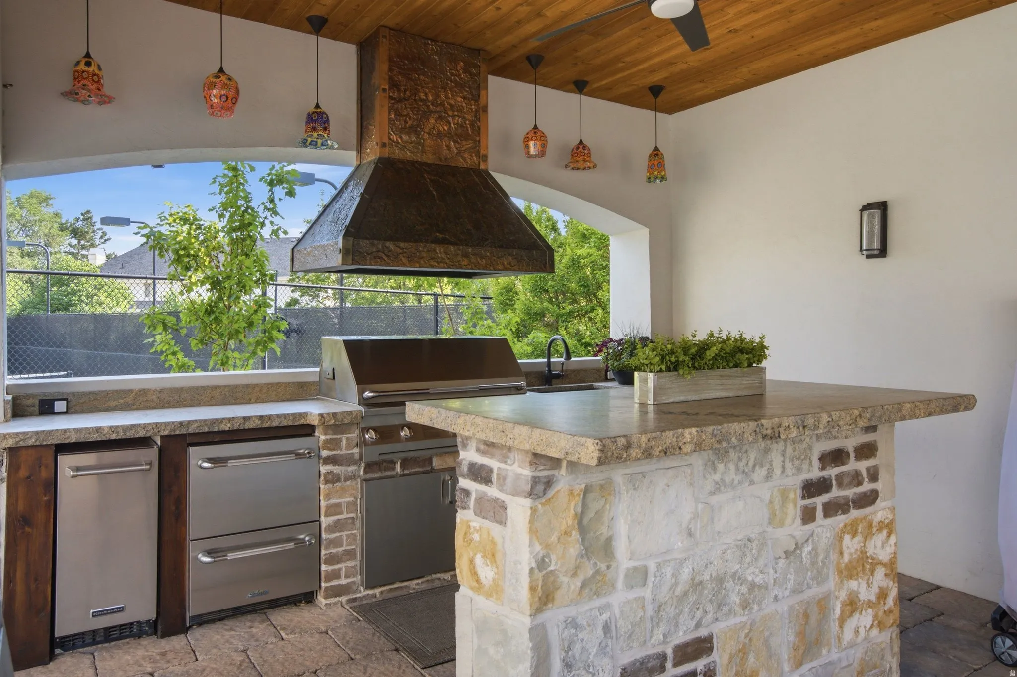 Outdoor kitchen featuring a stone and brick island, built-in grill, under-counter refrigerator, stainless steel drawers, and a wood-paneled ceiling with a fan