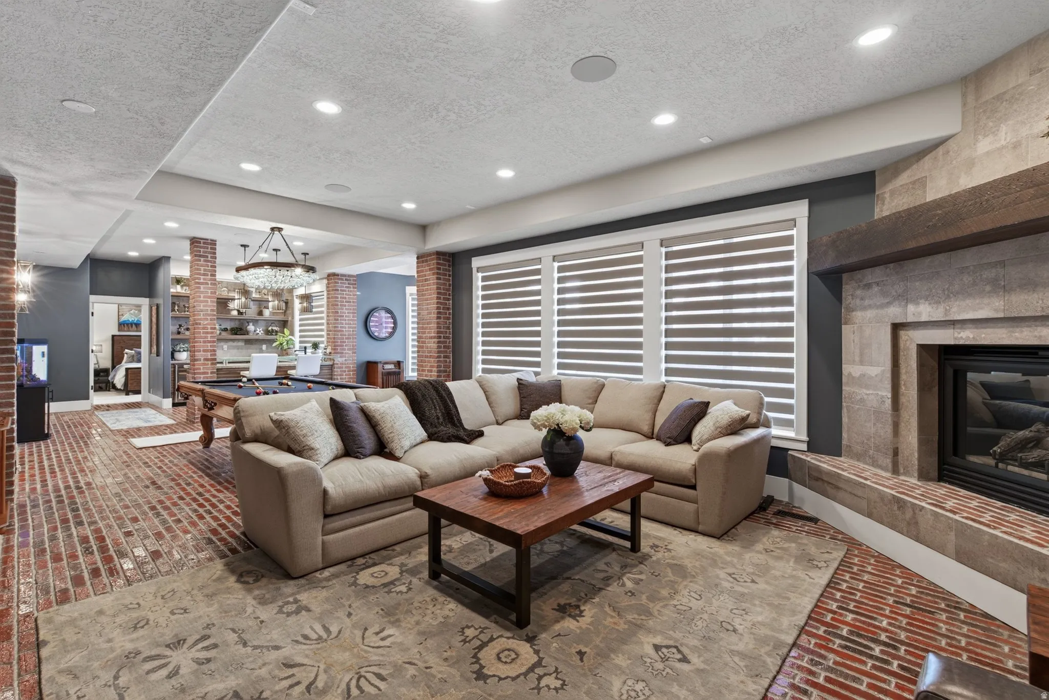 Open-concept living area with brick-finish flooring, recessed lighting, and a stone-clad fireplace featuring a wood mantel