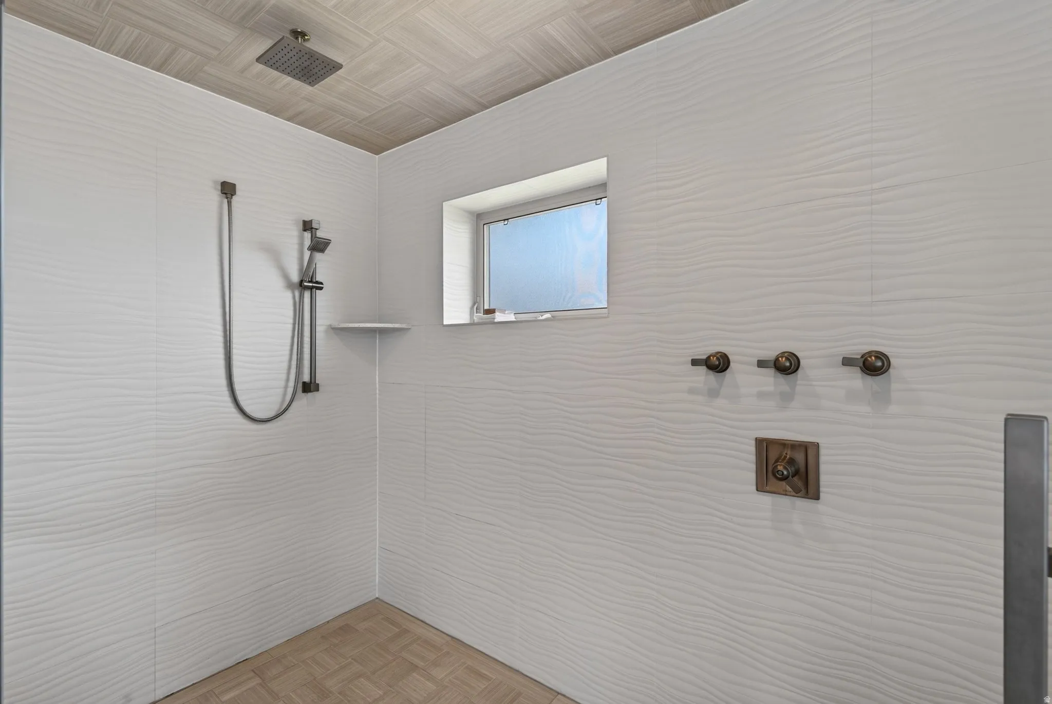Walk-in shower with textured wall tile, ceiling-mounted rainfall showerhead, handheld shower fixture, and a built-in shelf