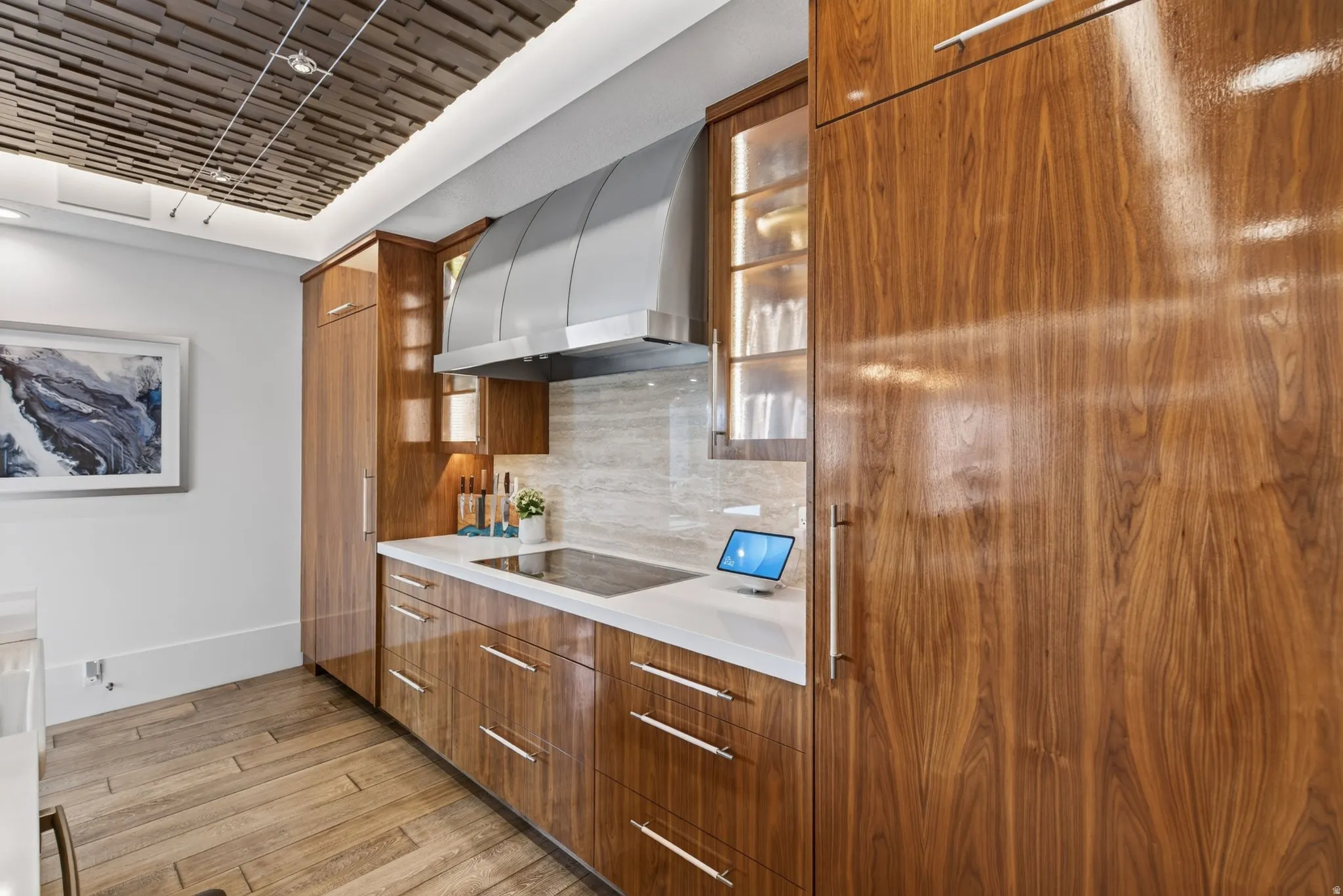 Glossy wood-finish cabinetry with linear hardware, light-toned countertop, integrated cooktop, stainless steel range hood, and wood-finish flooring