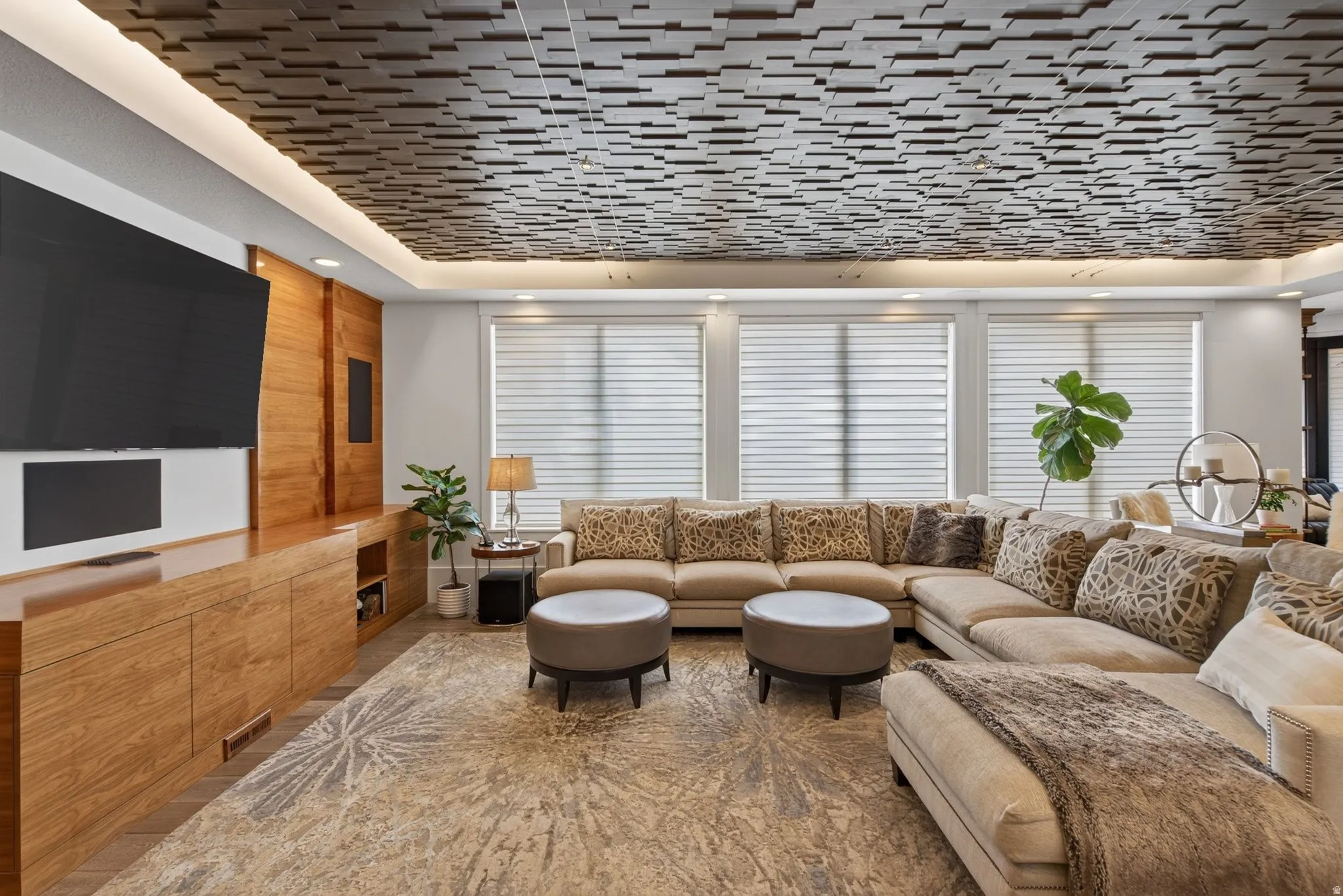 Coffered ceiling with recessed lighting, integrated wood-finish cabinetry, built-in media console, large windows with privacy blinds, and a patterned area rug