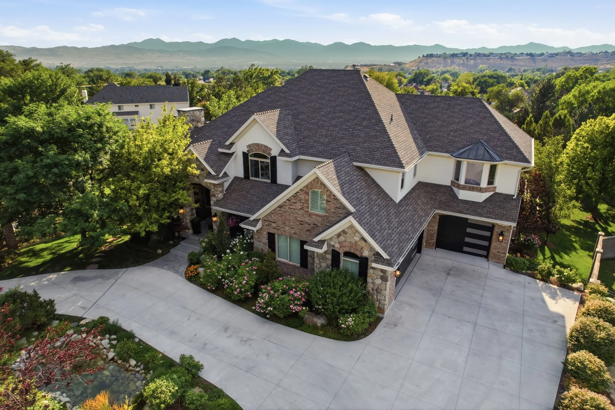 Stately exterior featuring stone and stucco finishes, an attached three-car garage, mature landscaping, and an expansive paved driveway