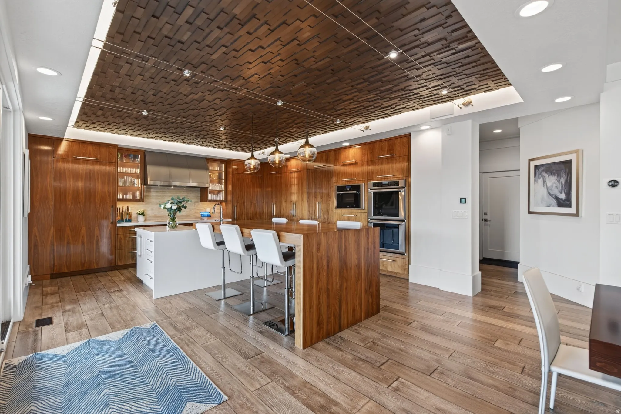 Hardwood flooring throughout, a white kitchen island with seating, built-in stainless steel double ovens, rich wood-finish cabinetry, and a distinctive wood-panel ceiling with integrated lighting