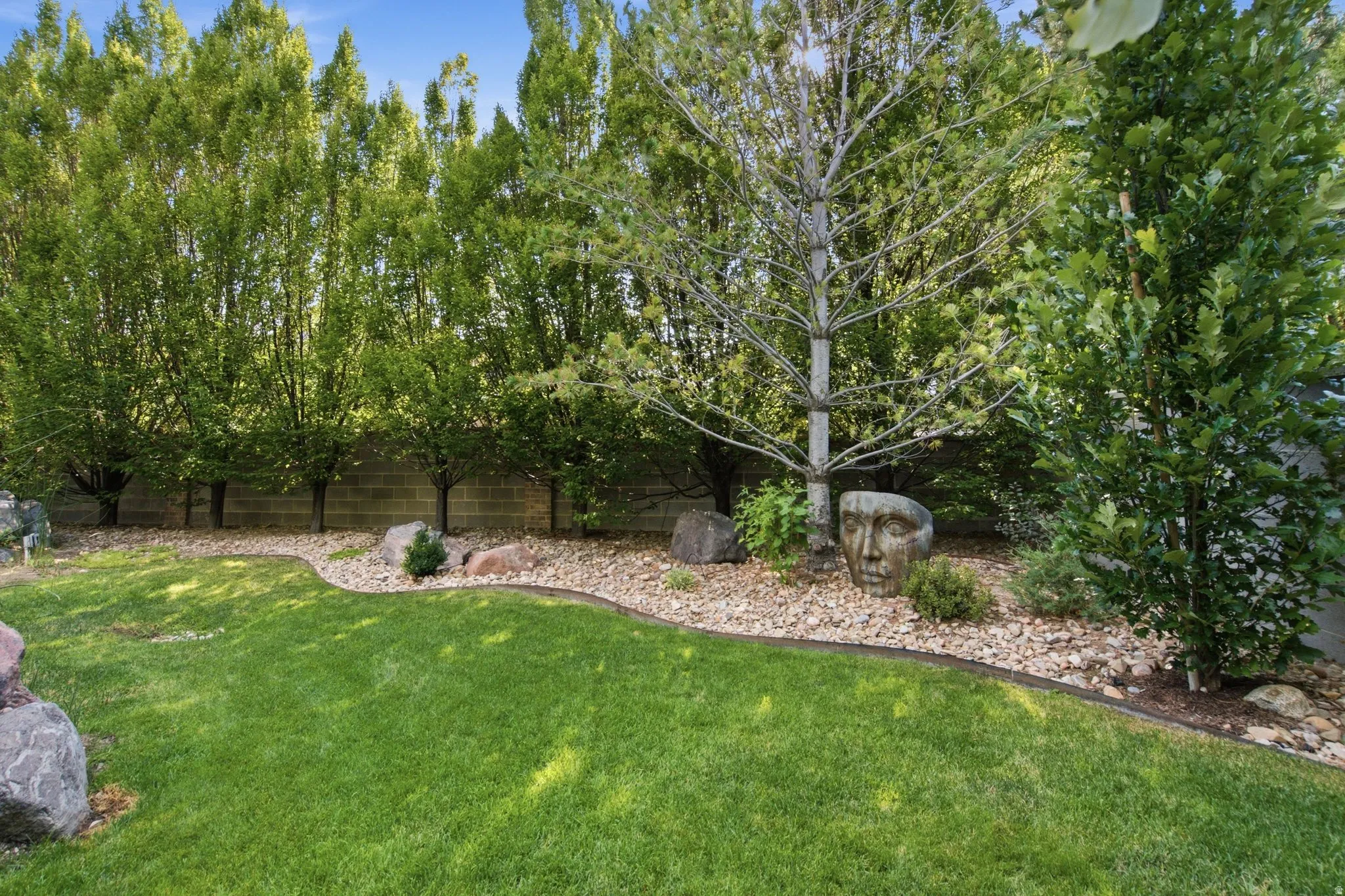 Manicured lawn with a curved landscape bed featuring decorative rocks, mature trees, and a concrete block retaining wall