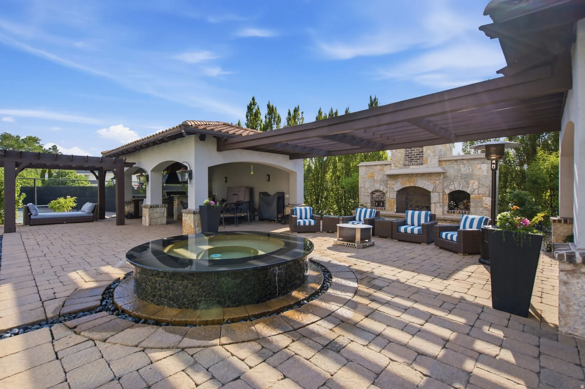 Expansive paver patio featuring a stone fireplace, wood pergola, and an outdoor kitchen with an arched entry