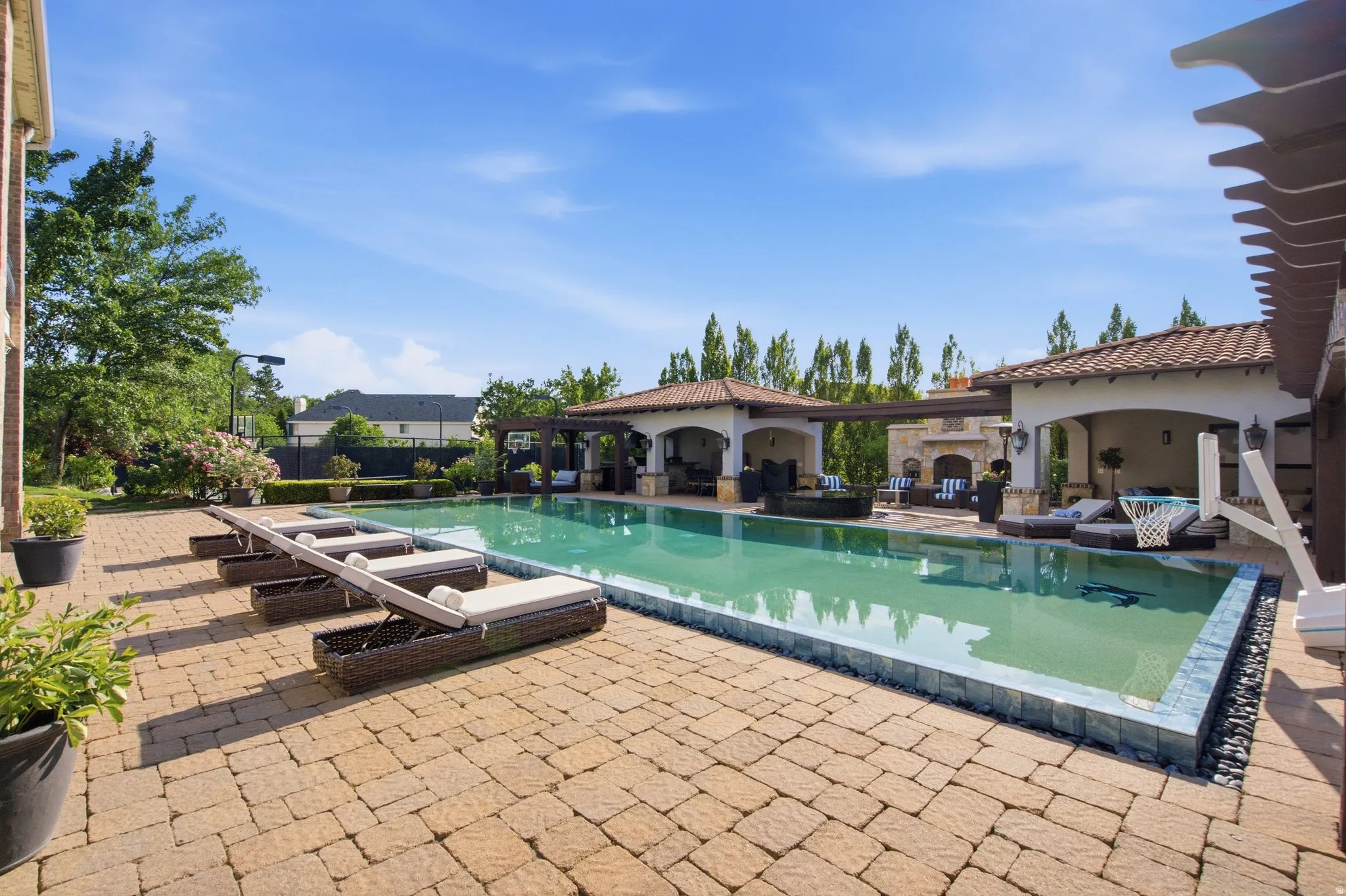 Rectangular swimming pool with dark tile surround, paver patio, two covered cabanas, and an outdoor fireplace