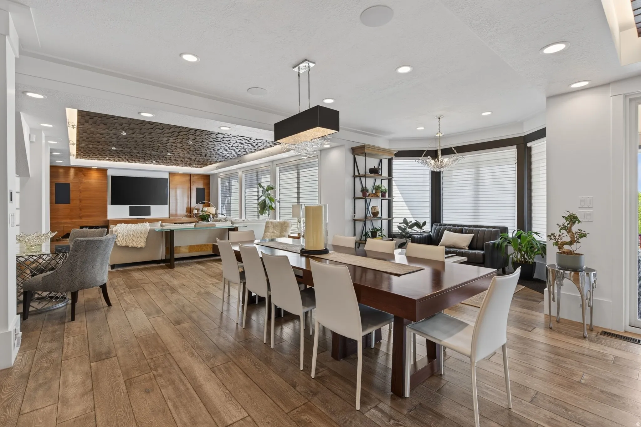 Expansive open-concept living area featuring wood-finish flooring, recessed lighting, a bay window with plantation shutters, and a distinctive textured ceiling accent