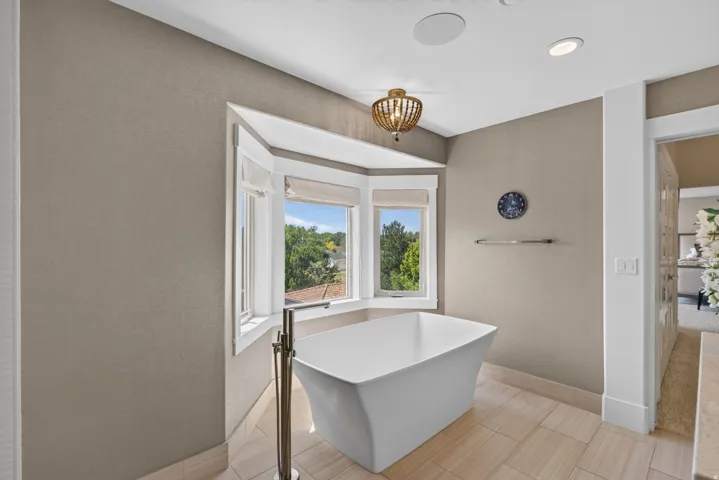 Freestanding soaking tub with floor-mounted brushed nickel faucet, bay window with window treatments, recessed lighting, and large format tile flooring
