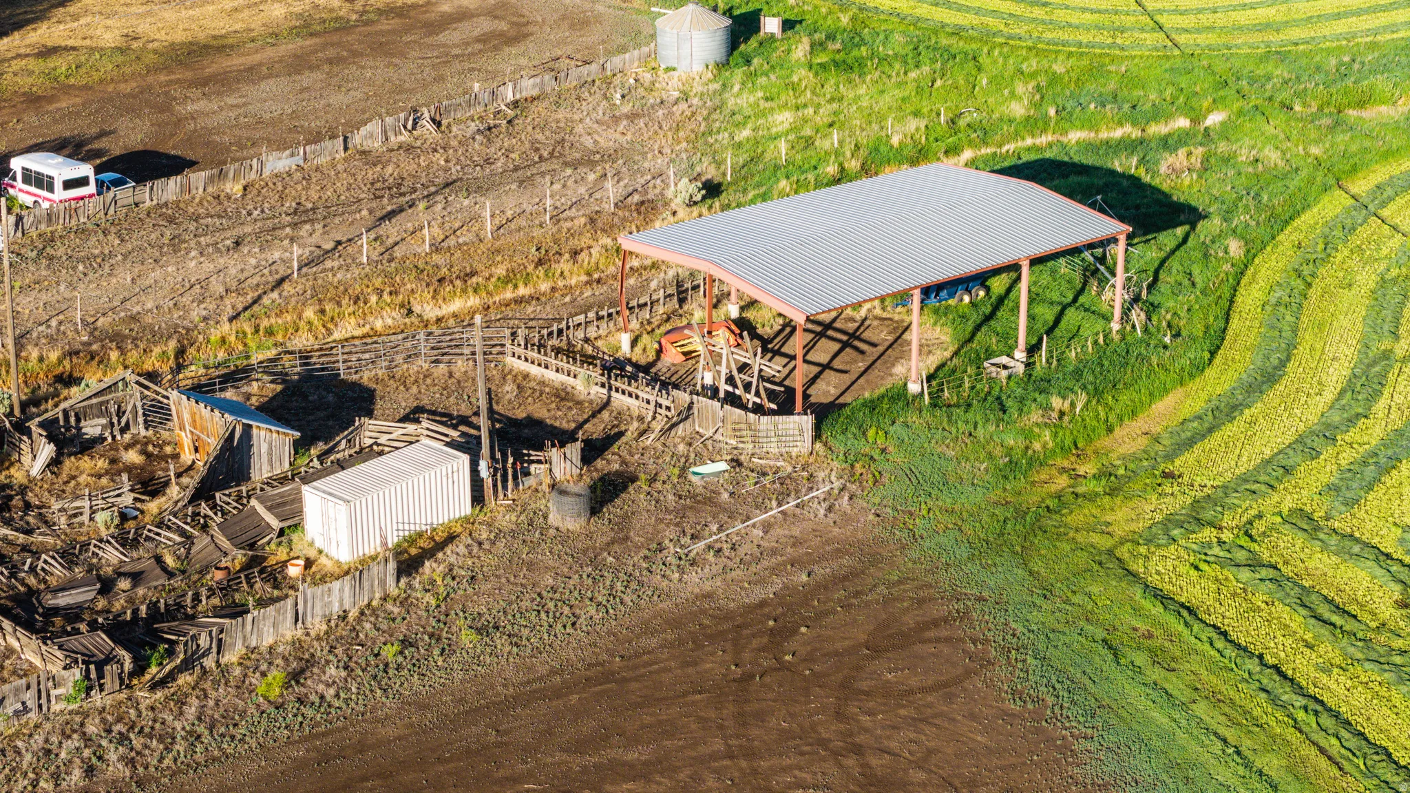 Corrugated metal shelter with open sides, surrounded by fenced enclosures and an adjacent cultivated field