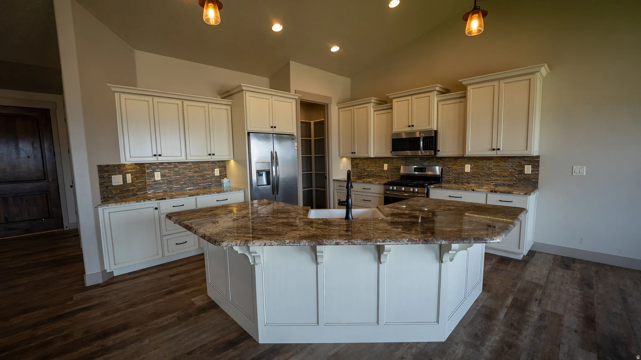 Spacious kitchen featuring an expansive center island, warm wood-finish flooring, white cabinetry, integrated stainless steel appliances, and a textured backsplash