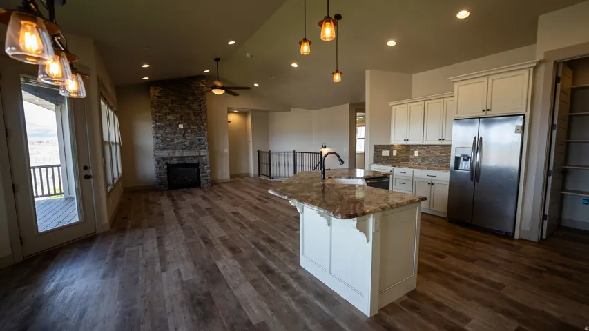 Vaulted ceiling with recessed lighting, stone-clad fireplace with mantel, wood-finish flooring, kitchen island with sink and faucet, and stainless steel refrigerator