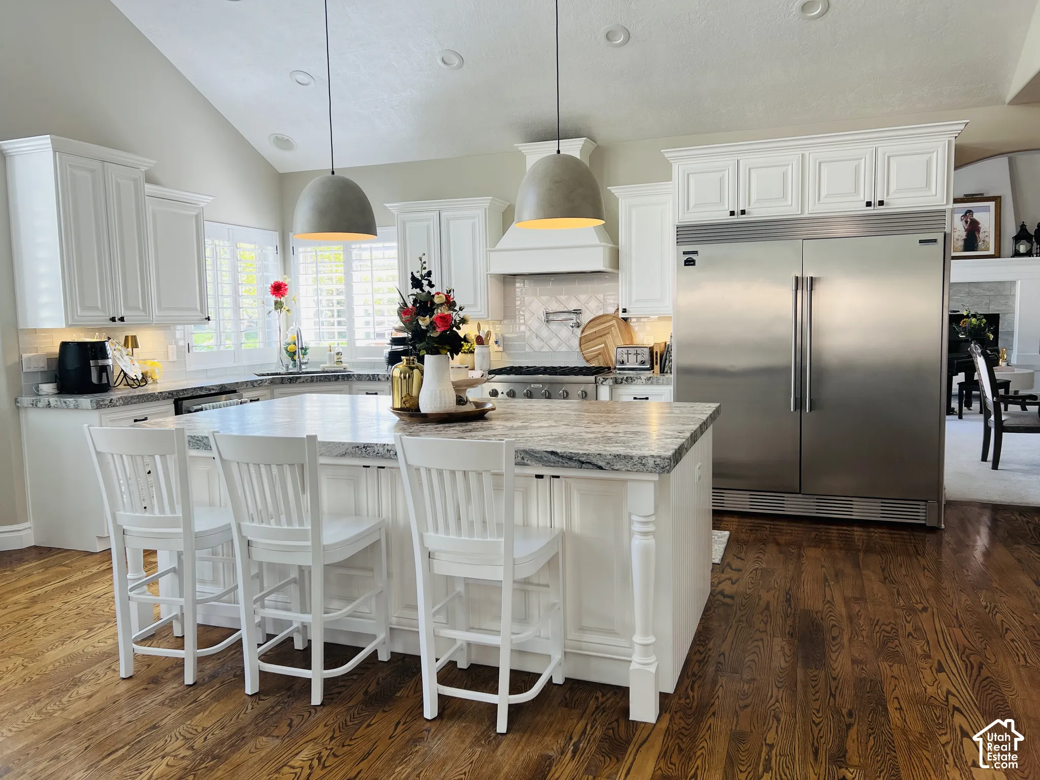 White cabinetry with crown molding, stainless steel refrigerator and range, large kitchen island with light-toned countertop, dark wood-finish flooring, and vaulted ceiling