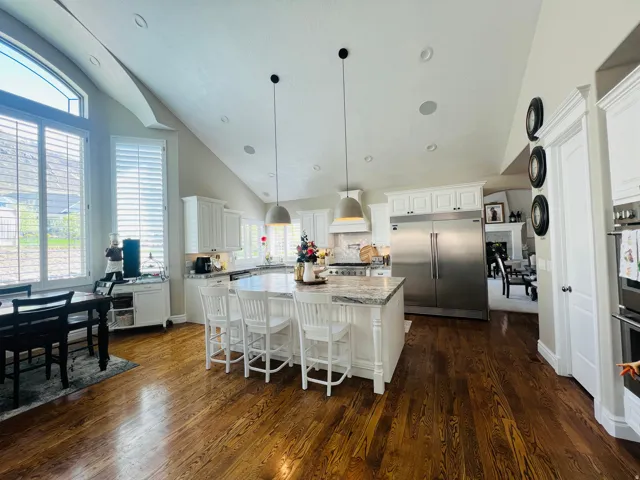 Vaulted ceilings, large windows with plantation shutters, wood-finish flooring, white cabinetry, and a center island with countertop seating