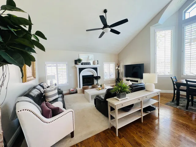 Vaulted ceiling with a ceiling fan, fireplace with a decorative mantel, plantation shutters, and wood-finish flooring