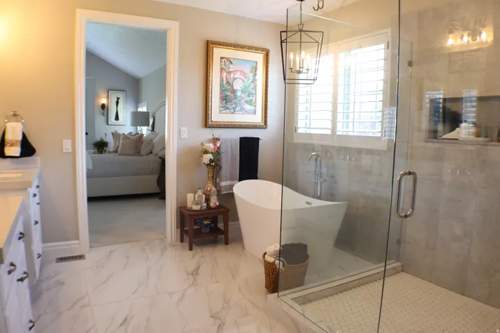 Freestanding soaking tub with floor-mounted faucet, glass-enclosed shower featuring a mosaic tile pan, shuttered window, and large format marble-look tile flooring