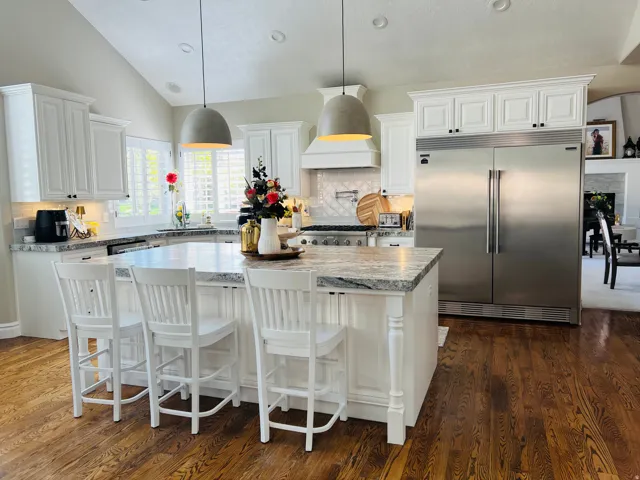 White cabinetry with crown molding, stainless steel refrigerator and range, large kitchen island with light-toned countertop, dark wood-finish flooring, and vaulted ceiling