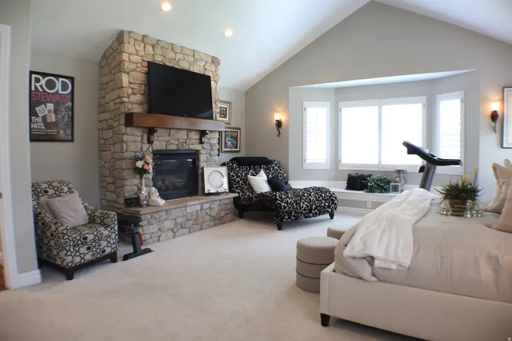 Stone-clad fireplace with a wood mantel, vaulted ceiling, bay window with built-in seating, and wall-mounted sconces