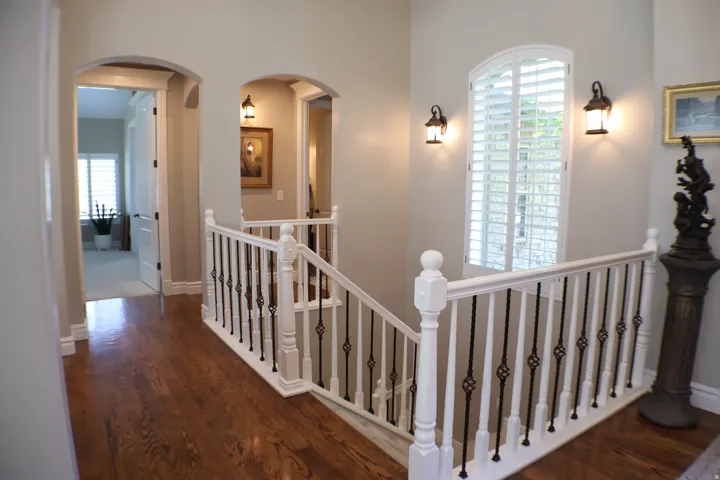 Hardwood flooring with a polished finish, elegant white newel posts, decorative wrought iron balusters, arched doorways, and a window featuring plantation shutters
