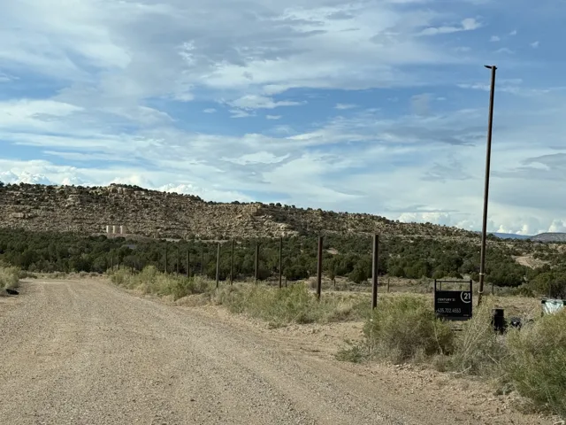 Gravel road entrance with a pole fence line, featuring dense foliage and a rocky hillside