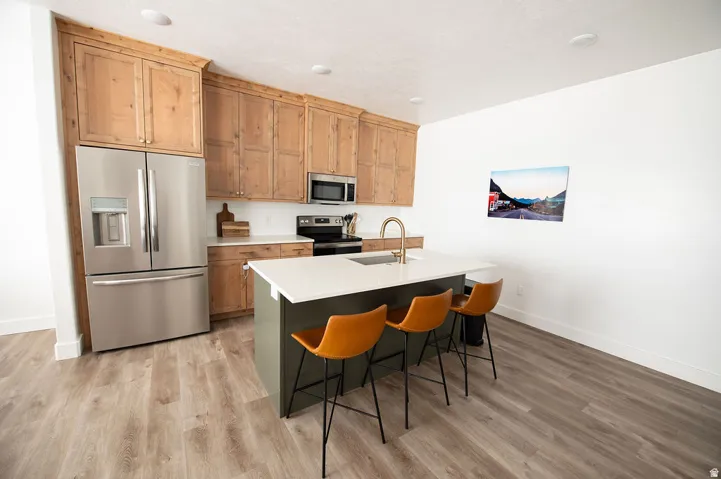 Kitchen featuring wood cabinetry, stainless steel appliances, a center island with a sink, white countertops, and wood-finish flooring