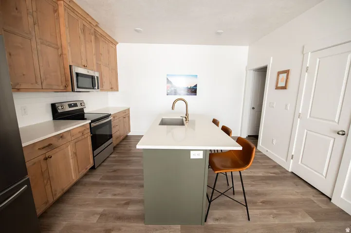 Kitchen featuring wood-finish cabinetry, stainless steel appliances, a subway tile backsplash, light-toned countertops, and a center island with a sink and brass-finish faucet
