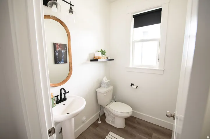 Pedestal sink with matte black faucet, oval wood-frame mirror, single window with roller shade, white toilet, and wood-finish flooring