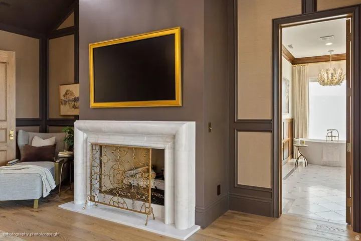 Sophisticated open-concept space featuring a two-sided fireplace with marble surround and decorative brass fireplace tools, illuminated flat-screen television with gold-leaf frame, hardwood flooring, and seamless access to adjoining rooms with dark wood t