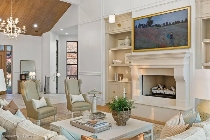 Vaulted wood-paneled ceiling, white chandelier, stone fireplace with mantel, built-in shelving, and large multi-pane windows