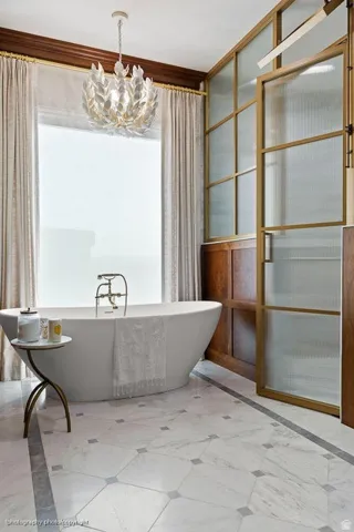 Freestanding soaking tub with brass fixtures, marble-patterned floor tiling, decorative chandelier, and frosted glass partition with brass framing
