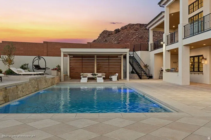 Rectangular swimming pool, stone-clad retaining wall, paved pool deck, covered outdoor living area with slatted wood wall, and multi-story exterior with balconies