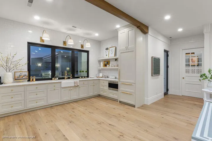 Light wood-finish flooring, white subway tile backsplash, white shaker cabinetry with gold-tone hardware, large black-framed window with a farmhouse sink, and a built-in microwave